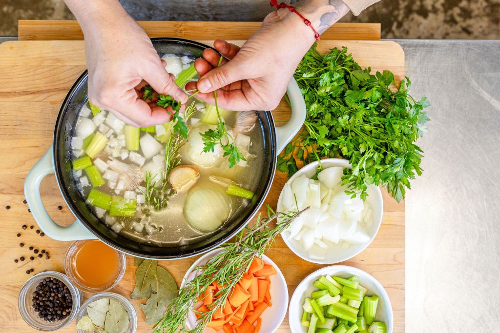 Chef’s hands preparing mirepoix beside a pot of simmering broth with fresh herbs.