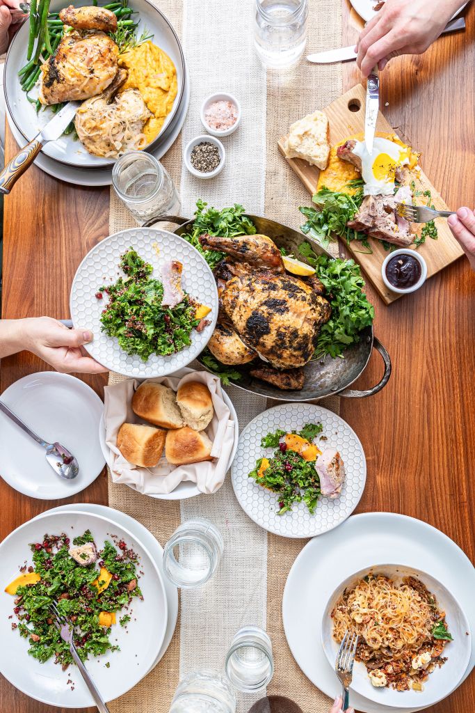 Guests gathered around a table sharing chef-prepared dishes, fresh salads, roasted chicken, house-made rolls, and seasonal fare during a communal dining experience